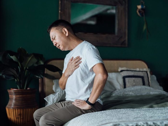 Senior Asian Man With Eyes Closed Holding His Chest In Discomfort, Suffering From Chest Pain While Sitting On Bed At Home. Elderly And Health Issues Concept