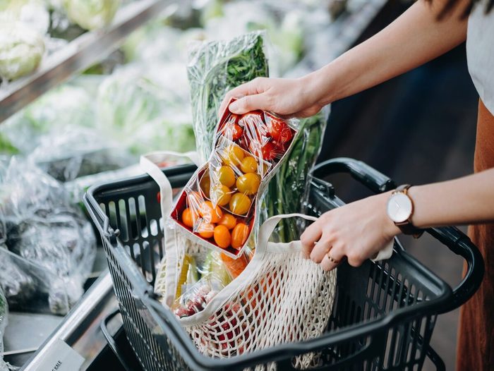 Cropped Shot Of Young Asian Woman Shopping For Fresh Organic Groceries In Supermarket And Putting A Variety Of Fruits And Vegetables Into Cotton Mesh Eco Bag In A Shopping Cart. Zero Waste Concept