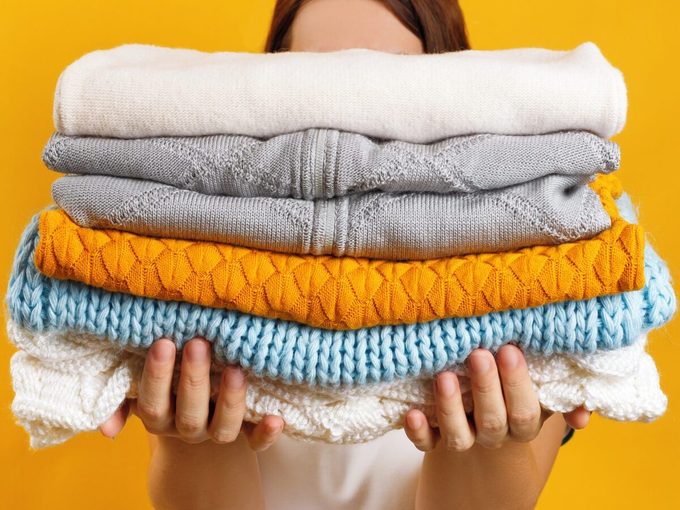 A Woman Holds A Stack Of Warm Knitted Clothes On A Background Of A Yellow Wall
