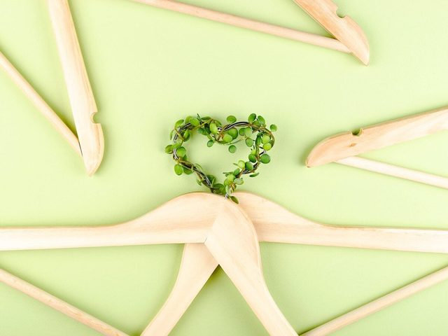 Conscious Consumption Slow Fashion Concept. Heart Of Clothes Hangers Entwined With Plant On Green Background.