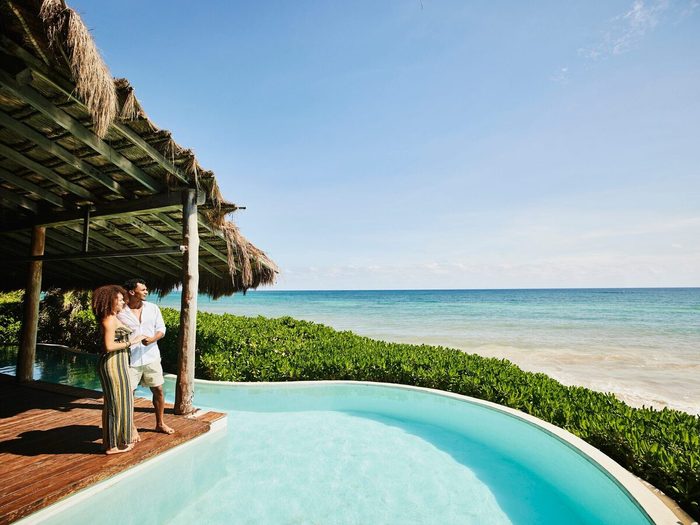 Wide Shot Of Couple Standing Poolside At Luxury Tropical Beachfront Villa Looking At View