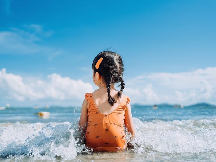 Rear View Of Lovely Little Asian Girl Sitting By The Seashore At The Beach And Being Splashed By Waves. Having Fun At Beach On A Sunny Summer Day